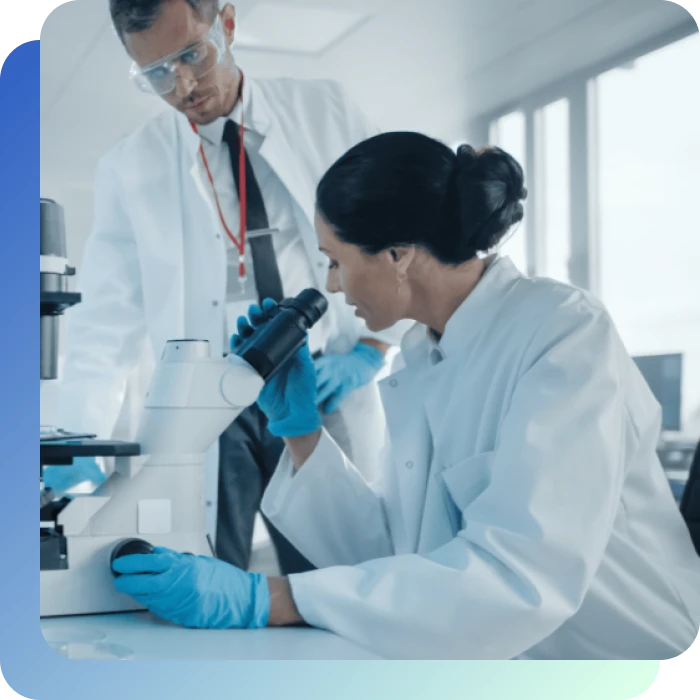 Eine Wissenschaftlerin arbeitet am Mikroskop ein Wisssenschaftler steht daneben und zeigt auf etwas.